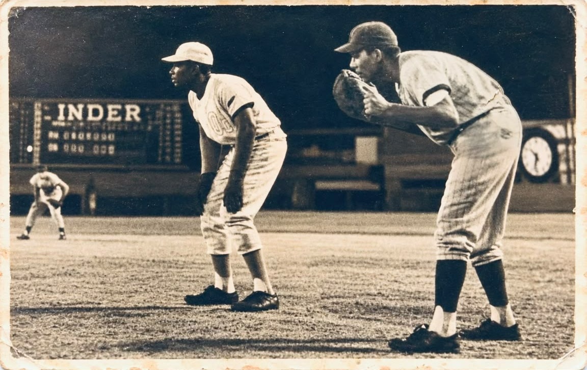 Dos peloteros en posición durante un juego nocturno, con el marcador del INDER al fondo
